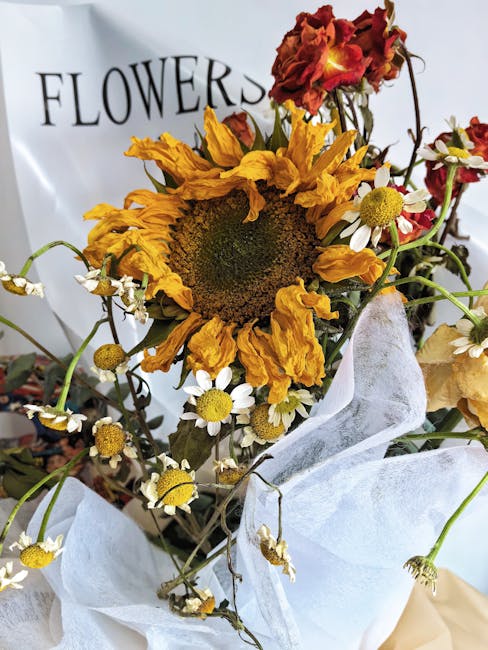 A close-up of a mixed floral arrangement featuring a prominent withered sunflower with large, drooping yellow petals and a dark central disk, surrounded by small white daisies with yellow centers, and