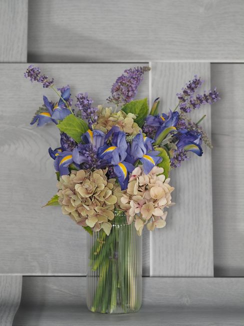 A floral arrangement in a clear glass vase featuring a mix of fresh flowers including purple irises with yellow accents, pale pink hydrangeas, light purple lavender sprigs, and green foliage. The bouq