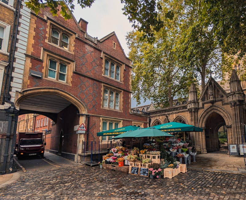 A flower stall situated on a cobblestone street near a historic brick building with an arched gateway, featuring an array of fresh flowers including roses, peonies, and tulips in vibrant pinks, whites